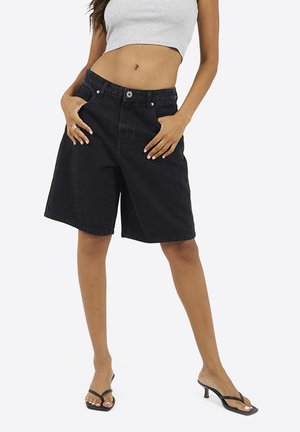 Kobieta ubrana w czarne, denimowe spodenki do kolan, szary, krótki tank top i czarne sandały na obcasie, stojąca z rękami w kieszeniach.