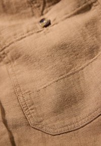 Tan bomullstyg med herringbone-mönster, med en bröstficka och en knappdetalj vid sömmen, som betonar textur och sömnad.