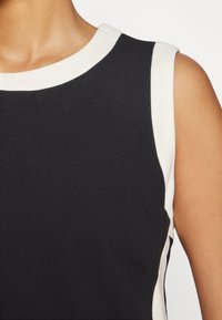 Haut noir sans manches avec bordure blanche autour du col et des emmanchures porté par une personne sur un fond blanc.