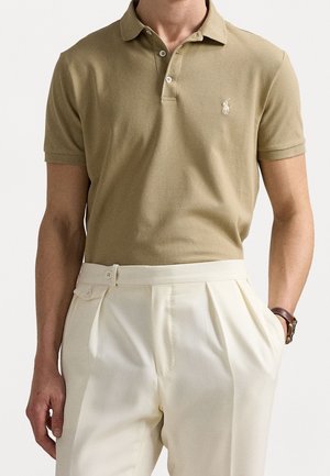 Homme portant un polo beige à manches courtes rentré dans un pantalon plissé crème taille haute avec une montre au poignet gauche.