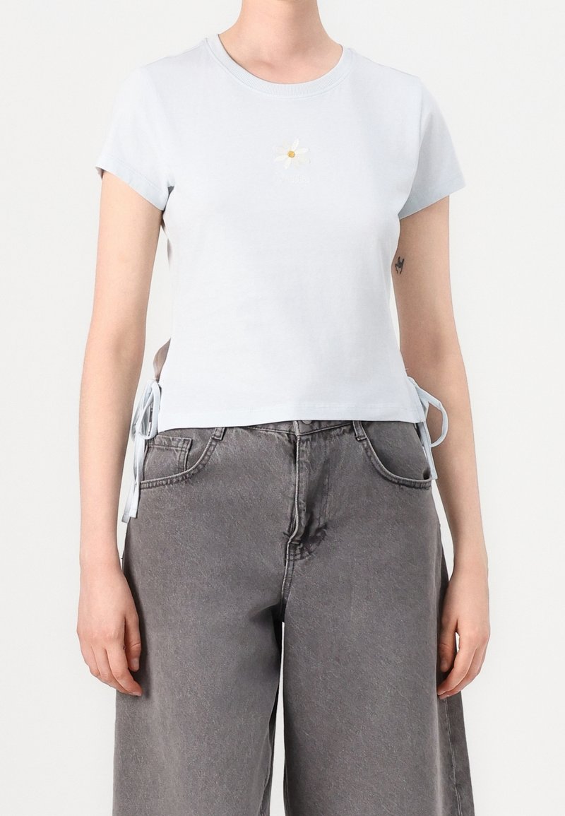 Personne portant un t-shirt court blanc avec un petit motif marguerite et des liens latéraux, associé à un jean gris ample taille haute.