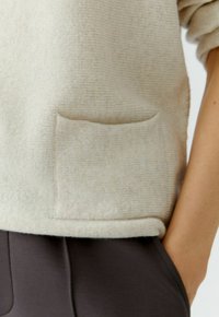 Helle beige Strickjacke mit strukturierter Oberfläche, ausgestattet mit einer Fronttasche und einem gerippten Saum, kombiniert mit dunkelgrauen Hosen.