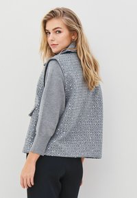 Blonde woman turned slightly back, wearing a gray long-sleeve shirt and a sleeveless gray sequin-embellished vest over black pants.