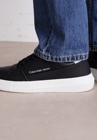 Sneaker in pelle nera con suola in gomma bianca, caratterizzata da una texture liscia, design con lacci e logo "Calvin Klein Jeans" sul lato.