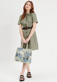 Olivenfarbendes Hemdkleid mit kurzen Ärmeln und einem schwarzen Gürtel mit runder Schnalle. Das Model hält eine Tasche mit floralen Mustern. Schwarze Sandalen.