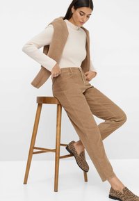Femme ajustant un pantalon en velours côtelé beige taille haute tout en étant assise sur un tabouret en bois, portant un pull côtelé crème et des mocassins à imprimé léopard.