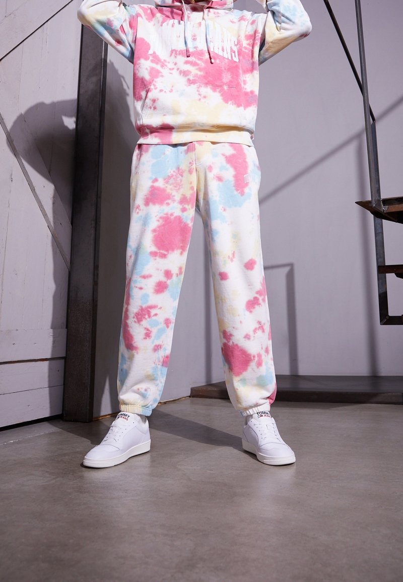Personne portant un sweat à capuche tie-dye pastel et un pantalon de survêtement assorti avec des baskets blanches, debout à l'intérieur près d'un escalier et d'un mur.