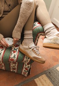 Person in beige leggings, cozy knit sweater, and beige sneakers sits on floral patterned cushioned chair holding a small floral teacup.
