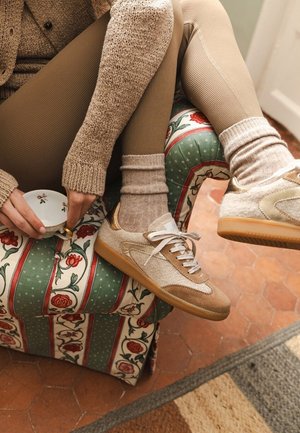 Persona in leggings beige, maglione in maglia morbida e sneakers beige è seduta su una sedia imbottita con motivo floreale, mentre tiene in mano una piccola tazza da tè floreale.