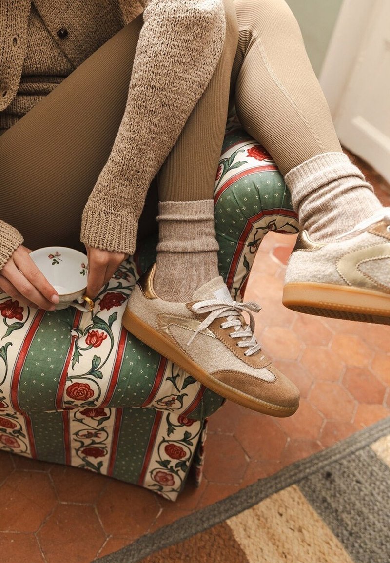 Une personne en leggings beige, un pull en tricot confortable et des baskets beiges est assise sur une chaise rembourrée à motif floral, tenant une petite tasse à thé florale.