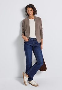 Blazer en tricot marron sur un haut crème, associé à un jean bleu et des baskets blanches. Le blazer présente un col rond et un seul bouton.