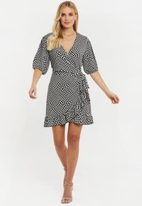 Vestido envolvente blanco y negro con lunares, mangas cortas abullonadas, bajo con volantes y un lazo en la cintura, hecho de una tela ligera.