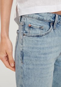 Jasnoniebieskie dżinsy mają dopasowany krój, pięć kieszeni oraz miedziane akcenty sprzętowe. Tkanina ma subtelnie wyblakłą fakturę.