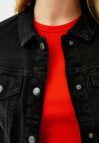 Giubbotto di denim nero con bottoni a pressione argento, indossato sopra una maglia rossa a costine. Primo piano focalizzato sulla texture del tessuto e sui dettagli dei bottoni.