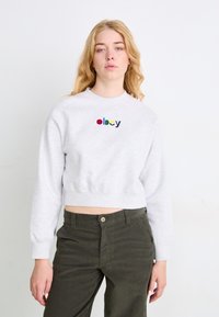 Sudadera gris corta con "obey" bordado en letras multicolores. Manga larga y cuello redondo. Combinada con pantalones de pana verde oscuro.