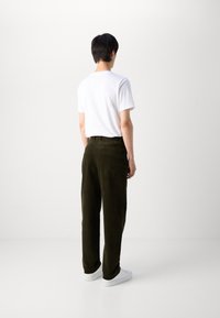 Zielone spodnie w kord do relaksowanego kroju, w połączeniu z białą koszulką z krótkim rękawem. Model stoi tyłem, prezentując gładką fakturę materiału.