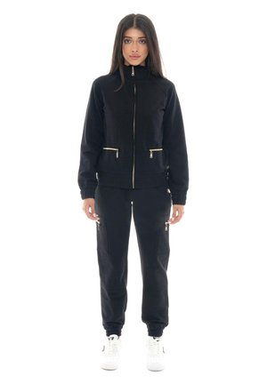 Une femme porte une veste noire zippée assortie à un pantalon de jogging cargo doté de fermetures éclair dorées, ainsi que des baskets blanches, sur fond blanc.