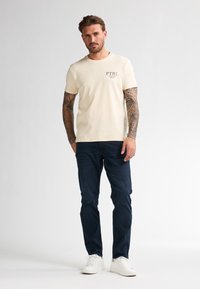 Svetle béžové bavlněné tričko s centrálně umístěným potištěným logem v tmavě hnědé barvě. Kombinováno s tmavě modrými džíny a bílými teniskami.