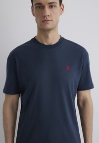 Marineblaue Baumwoll-T-Shirt mit klassischem Rundhalsausschnitt und kurzen Ärmeln, das ein kleines, rotes gesticktes Logo auf der Brust trägt.