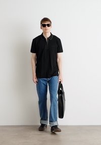 Polo nero con dettagli della zip al colletto, jeans blu, occhiali da sole neri e una borsa nera, in piedi contro uno sfondo bianco semplice.