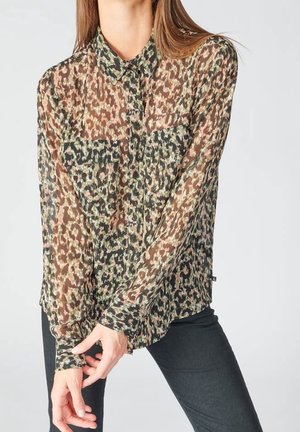 Kobieta w przezroczystej, długorękawowej bluzce w panterkę z zapinanymi na guziki kieszeniami, w czarnych dopasowanych spodniach, pozująca z złączonymi dłońmi.