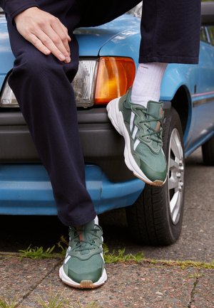 Person, die grüne und weiße Sneaker und dunkle Hosen trägt, sitzt vorne auf einem blauen Auto mit einem Fuß auf dem Boden.
