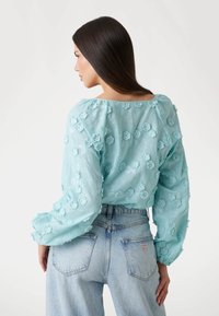 Lysblå gennemsigtig bluse med blomsterapplikationer, lange puffærmer og en bred halsudskæring, parret med højtaljede lysblå jeans.