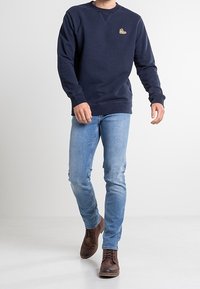 Marineblaue Sweatshirt mit einem kleinen Schuh-Logo auf der Brust, kombiniert mit hellblauen Jeans und braunen Schnürschuhen. Lässige Passform und Design.