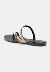 Beige mokka thong sandaalit, joissa on ohut musta hihna ja kultainen solki, ja jotka sisältävät littean tummanruskean pohjan.