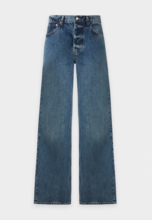 Femme aux longs cheveux foncés portant un haut sans manches bleu clair, un jean bleu à jambes larges et des chaussures noires, debout avec une main dans la poche.