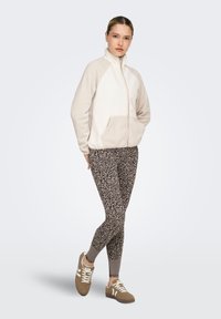 Veste en polaire avec des accents beiges, fermeture éclair à l'avant et poches, associée à des leggings à motifs dans des tons terreux et des baskets marron à lacets.