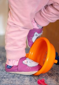 Fuchsia Leder-Babyschuhe mit einem Katzenmotiv, cremefarbenen Sohlen und elastischen Bündchen. Liegen neben einer orangefarbenen Spielschale auf einer strukturierten blauen Oberfläche.