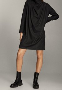 Robe noire drapée à manches longues, coupe décontractée, tissu texturé, tombant au-dessus du genou, associée à des bottines noires.