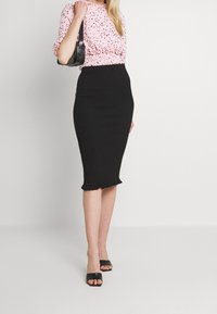 Blouse rose à motifs avec taille smockée, associée à une jupe crayon noire moulante avec un ourlet volants. Talons blocs ouverts noirs.