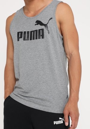 Mężczyzna ubrany w szarą koszulkę bez rękawów Puma i czarne krótkie spodenki Puma, stojący z jedną ręką w kieszeni na białym tle.