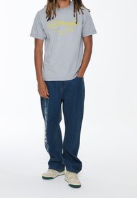 Lichtgrijs T-shirt met korte mouwen met een gele grafische print, gecombineerd met losse blauwe jeans met witte letters aan de zijkant en casual schoenen.