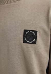 Camicia a maniche lunghe color khaki con una texture morbida, dotata di una patch quadrata nera con testo e logo bianchi sulla parte sinistra del petto.