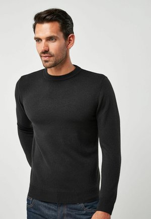 Suéter negro ajustado de manga larga con un cuello redondo acanalado, hecho de un material suave y texturizado. Lleva puesto sobre unos jeans de denim.