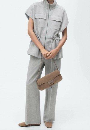 Vrouw draagt lichtgrijze broek met wijde pijpen en een kortärmelig ritstop met zakken aan de voorkant, houdt een bruine suède handtas vast en draagt bijpassende schoenen.