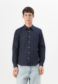 Joven con cabello corto castaño, vistiendo una camisa de botones azul marino oscuro y jeans azul claros, de pie contra un fondo blanco.