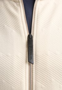 Beige gewatteerd jack met een gestructureerd zigzagpatroon. Ritssluiting met een zwarte trekker en merkdetail. De binnenkant toont een donkere stof.