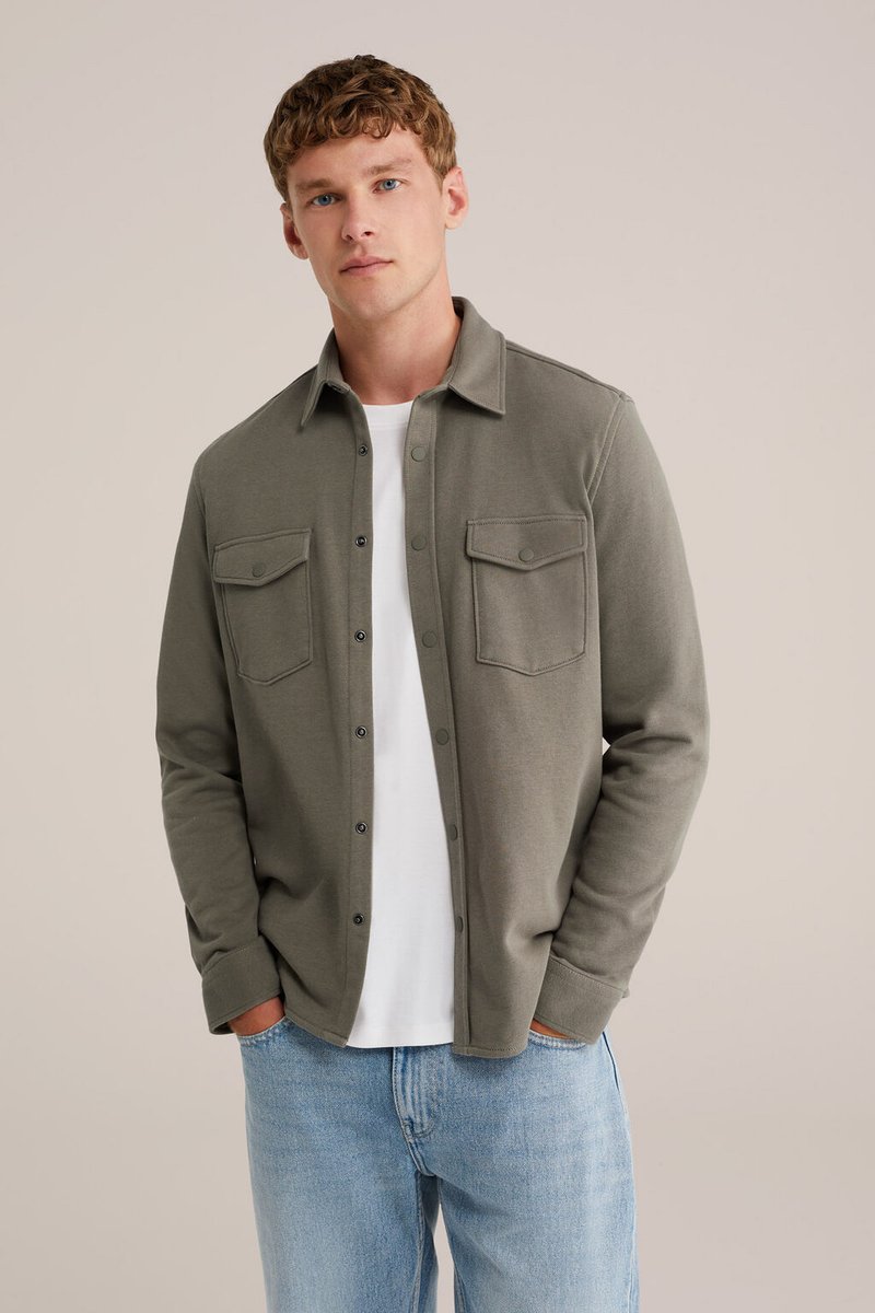 Joven con una chaqueta de camisa de color verde olivo sobre una camiseta blanca y jeans de color azul claro, de pie con las manos en los bolsillos.