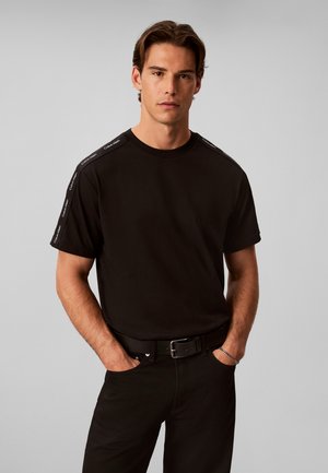 Άνδρας που φοράει μαύρο κοντομάνικο t-shirt με λογότυπα Calvin Klein στις ρίγες των ώμων, μαύρο παντελόνι, ζώνη, με τα χέρια στις τσέπες.