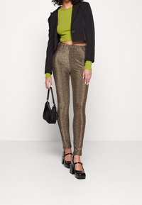 Kiiltävät kultaiset legginsit, joissa on joustava vyötärö, yhdistettynä mustaan lyhytvanteiseen bleiseriin ja vihreään pitkähiharaiseen paitaaan. Asusteina pieni musta laukku.