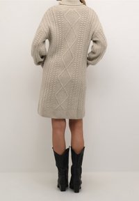 Personne portant une robe-pull beige en maille torsadée et des bottes en cuir noires mi-mollet, debout devant un fond uni.