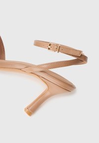 Bežové kožené sandále na kitten podpätku so štíhlym remienkom na okolo členka a zlatou sponou na bielom pozadí.