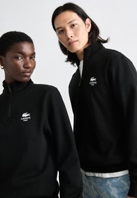 Juodas užtrauktukas megztinis su aukštu apykakle, balta Lacoste logotipu ir minkšta tekstūra. Du modeliai stovi arti, demonstruodami dizainą.