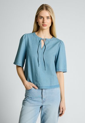 Kobieta ubrana w jasnoniebieski denimowy top z krótkim rękawem i wiązaniem przy dekolcie, w połączeniu z jasnoniebieskimi dżinsami, stojąca na jednolitym tle.