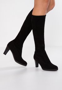 Bottes noires en daim qui dépassent le genou, à bout pointu et talon empilé, présentant une finition lisse et un détail de couture subtil.