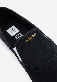 Svart slip-on Adidas sko med ovandel i mocka, guld Adidas-logga på tungan och vita elastiska sidodelar.